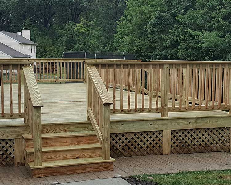 A wooden backyard deck with stairs, a railing, and lattice skirting, built over a paver patio next to a house.
