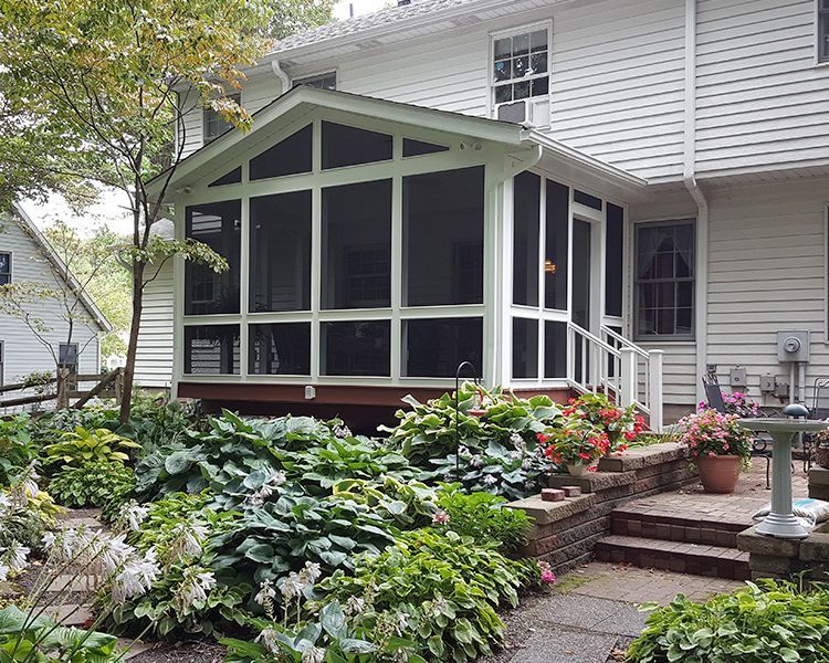 A white-sided house with a screen porch extension, overlooking a lush garden with stone stairs and potted flowers.