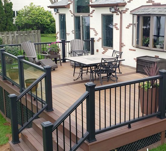 A tiered composite deck featuring a dining table, chairs, and black metal railings with glass panels.