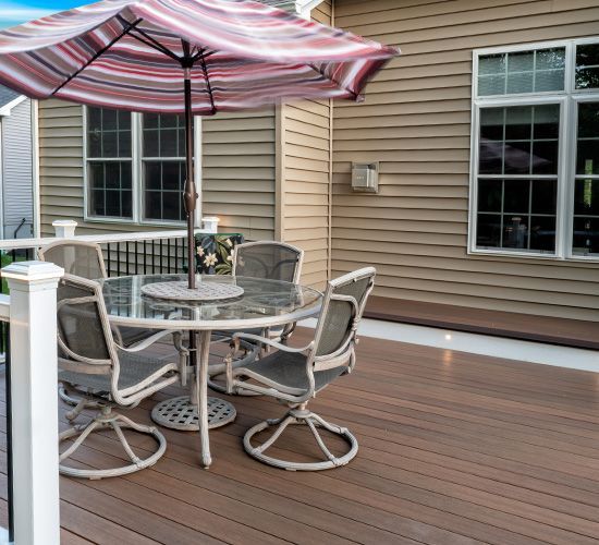 A patio set with a round glass table and four chairs under a striped umbrella on a brown composite deck.