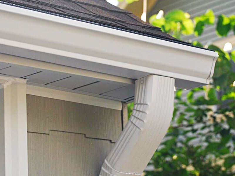 A white metal gutter and a vertical downspout attached to the corner of a house with tan siding and dark roof shingles.