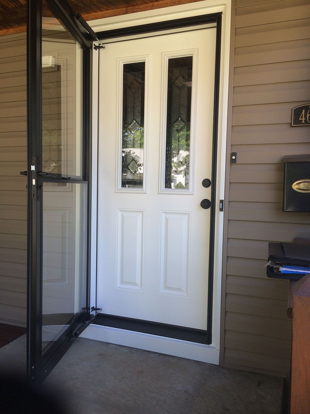A white front door with two vertical glass panels, framed by a dark screen door on a porch with beige siding.