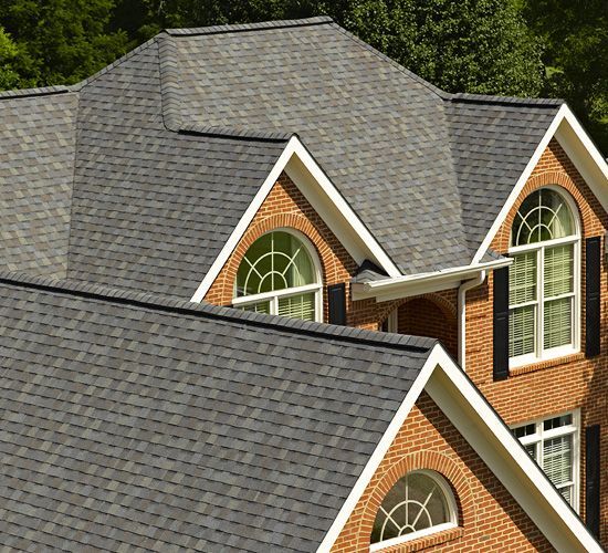 A close-up view of a multi-gabled roof with dark grey shingles on a red brick house featuring arched windows.