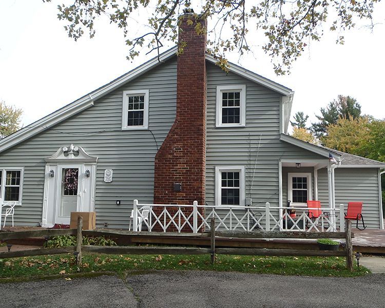 A light gray house with a brick chimney, a white lattice porch, and red chairs sits behind a wooden fence.