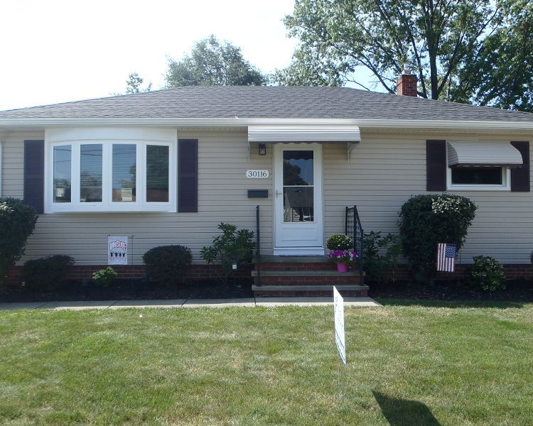 A single-story, light tan suburban house with dark shutters, a bay window, front stairs, and a well-manicured lawn.