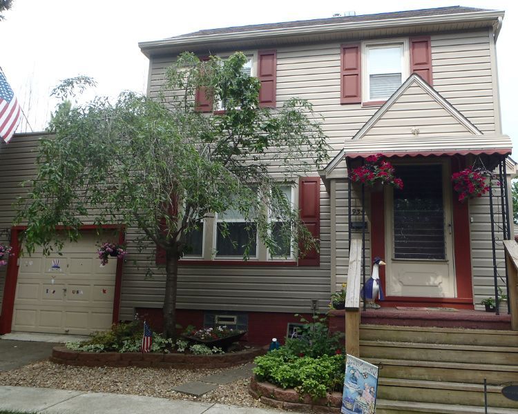 A two-story tan house with red shutters, a garage, a front porch with stairs, and a weeping tree in the front yard.