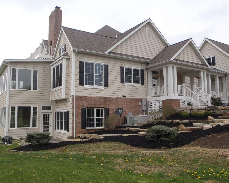 A multi-story beige house with brick accents, white trim, black shutters, and a covered porch on a landscaped lawn.