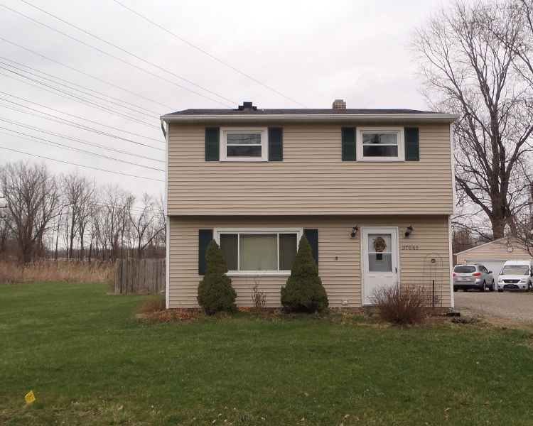 A two-story, beige vinyl-sided house with a white door, dark shutters, and a lawn under a cloudy sky.