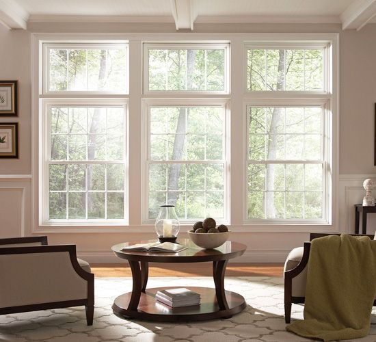 Three large white windows above a circular wooden coffee table with a bowl of spheres, between two chairs in a room.