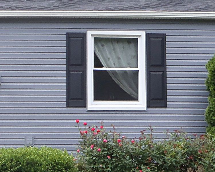 A single window with black shutters, white trim, and white curtains on a light blue-gray horizontal-sided house wall.