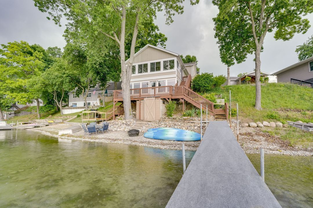 Lakeside house with wooden deck, dock, and kayak; trees and cloudy sky in background.