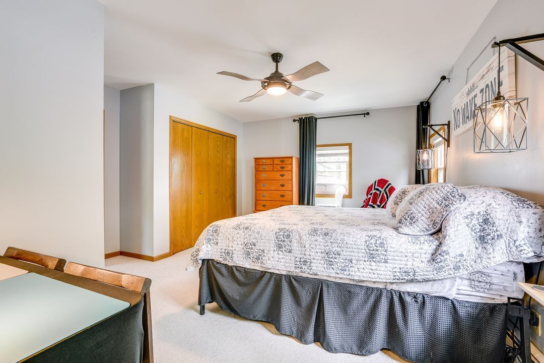 Bedroom with bed, dresser, wooden closet doors, and ceiling fan. Light gray walls, cream carpet, and dark bed skirt.
