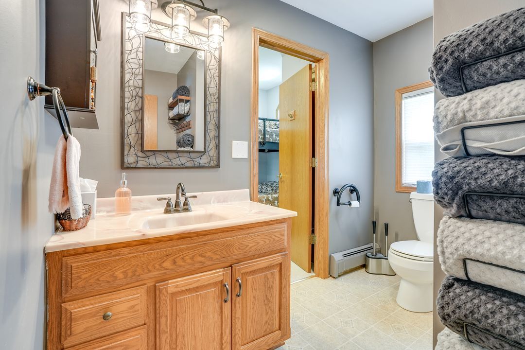 Bathroom with wooden vanity, mirror, toilet, stacked towels, and a doorway.
