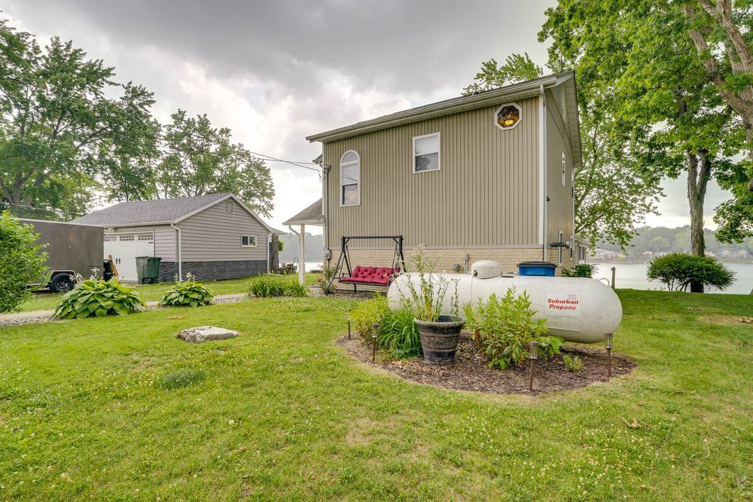 A house with a propane tank in front, next to a garage, on a grassy lawn near water. Overcast day.