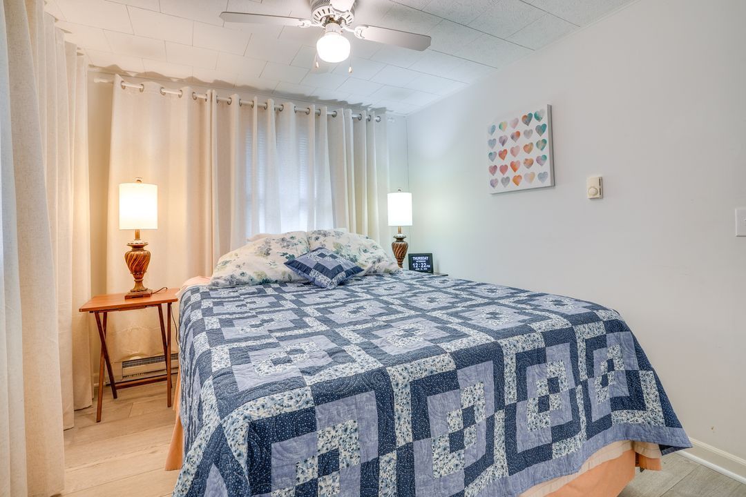 Bedroom with bed, blue quilt, white curtains, lamps, and a small side table.