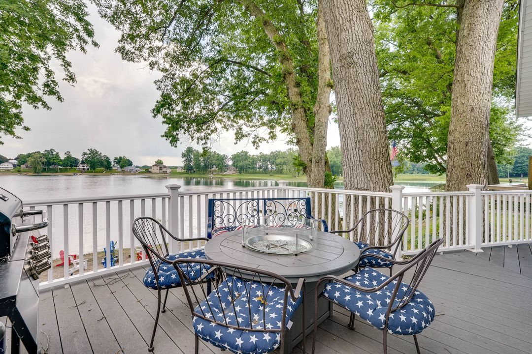 Patio with round table and chairs overlooking a lake. White railing, trees, and gray deck.