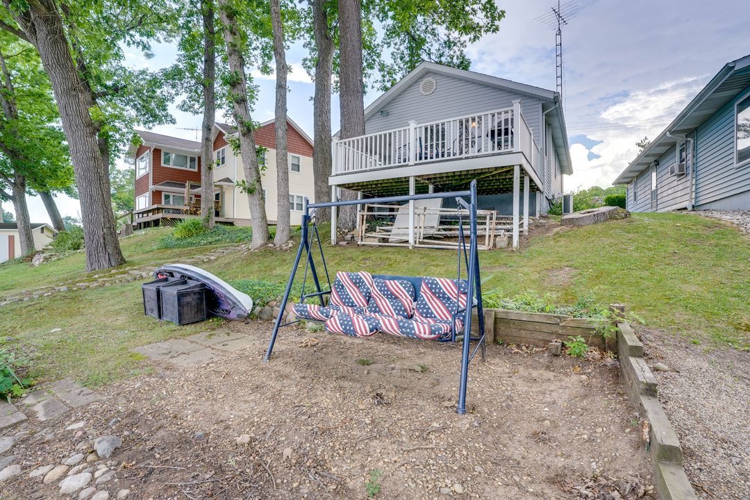 A swing set with American flag cushions sits in front of a gray house with a deck overlooking a body of water.