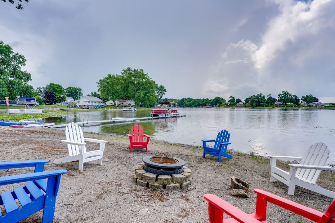 Lakeside scene with colorful Adirondack chairs around a fire pit. Boats and homes are in the background. Overcast sky.