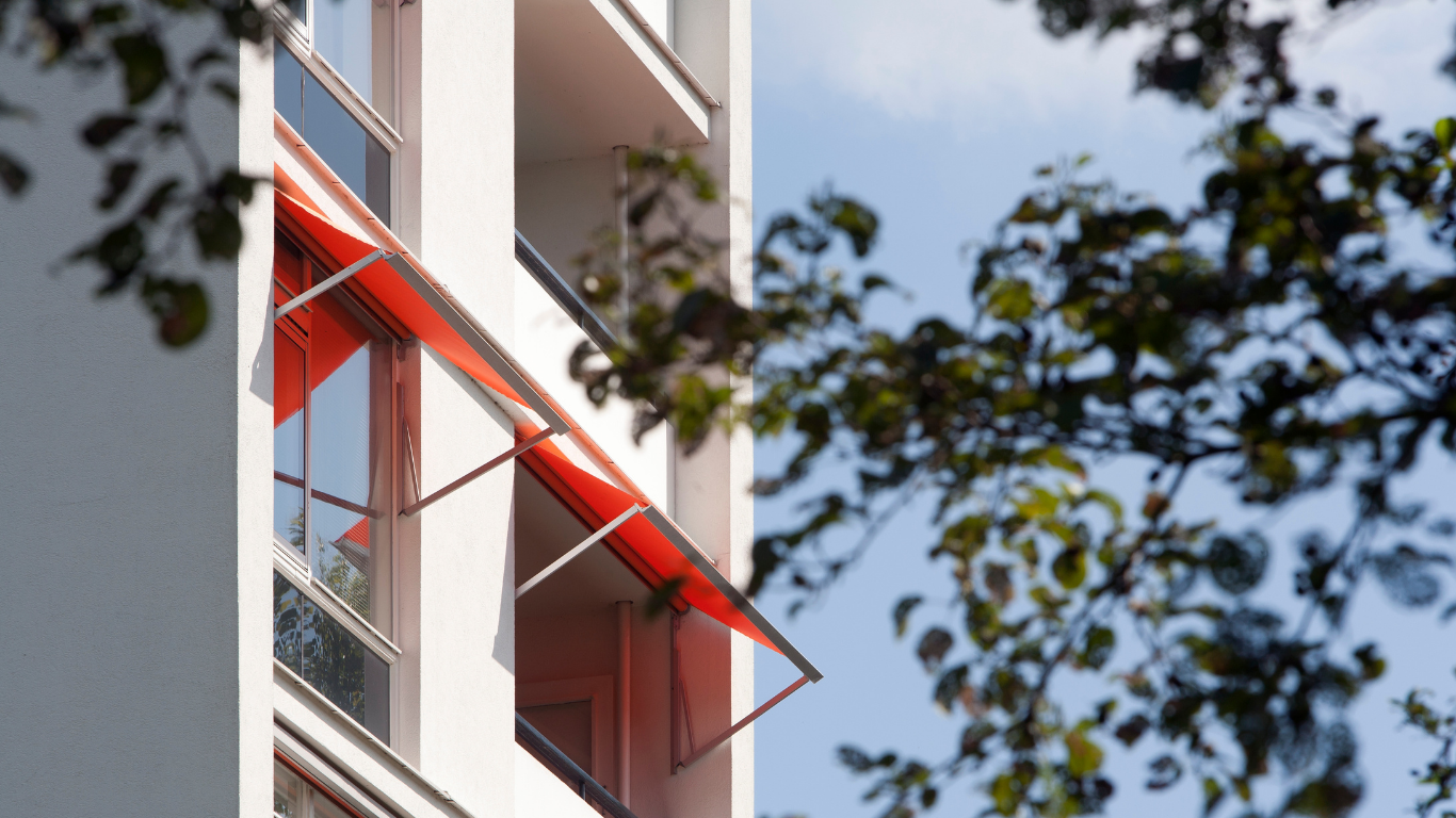 Red awnings on a white building, partially obscured by green tree leaves against a blue sky.