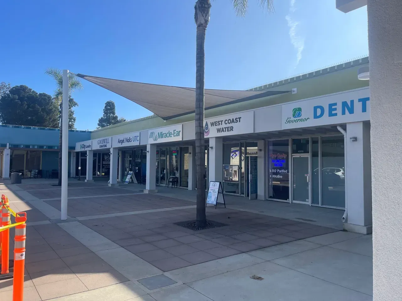 Shops under a canopy on a sunny day. A dental office, cafe, and other businesses are visible.