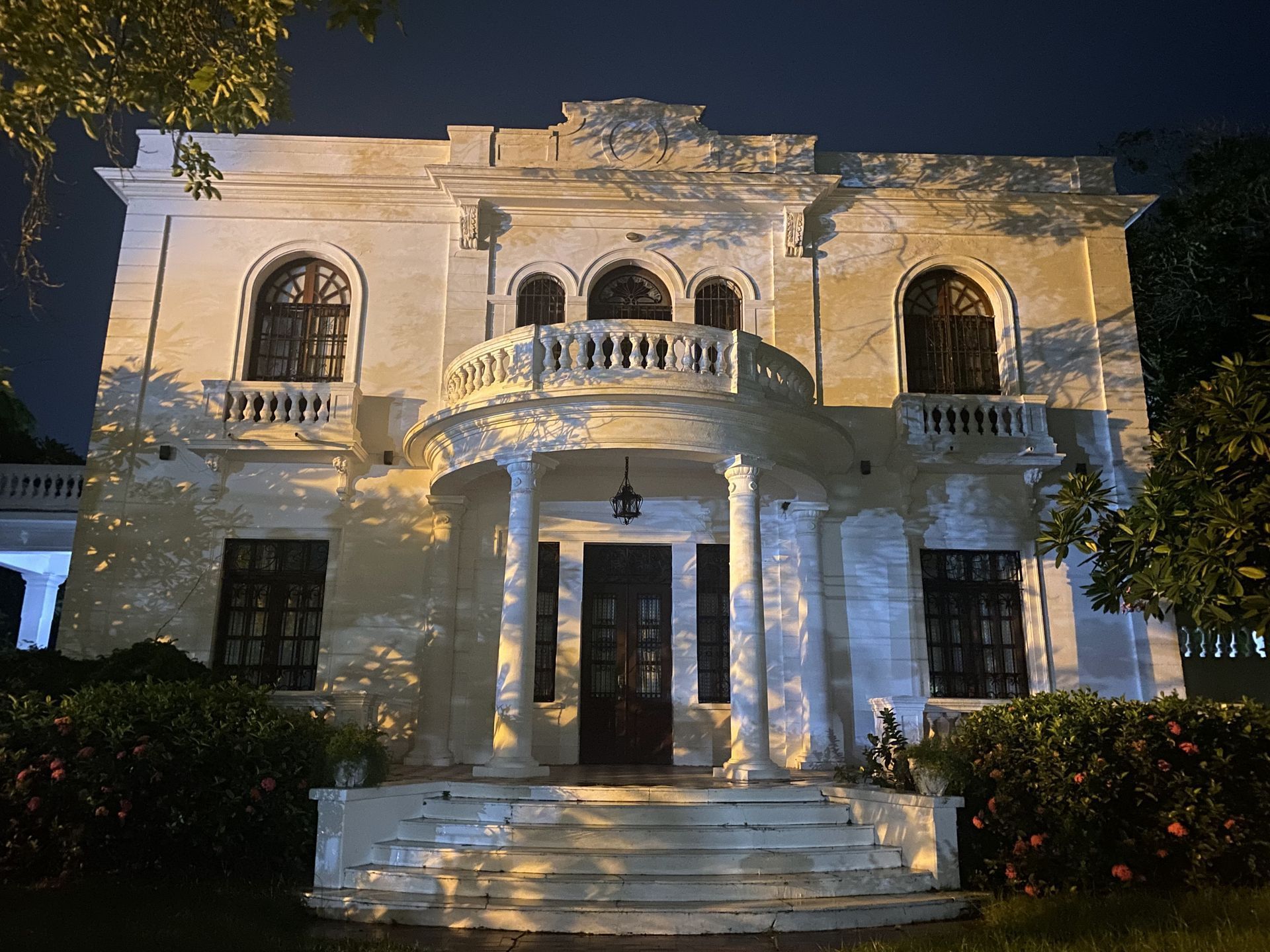 White two-story building with a balcony, columns, and arched windows, lit at night.