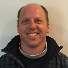 Man with short brown hair smiles, wearing a dark blue jacket against a white wall.