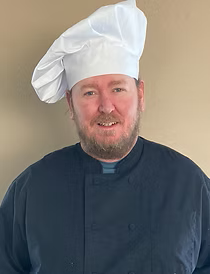 Chef wearing a white hat and black coat, smiling in front of a neutral-colored wall.