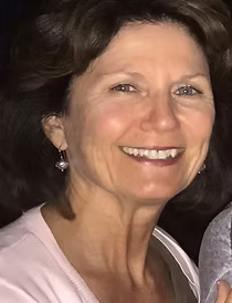 Woman with short brown hair smiles, wearing a light pink shirt, and pearl earrings.
