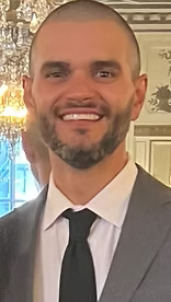 Man in a grey suit smiles, wearing a black tie and white shirt in a formal setting.