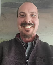 Man with a handlebar mustache smiles, wearing a gray shirt and dark jacket.