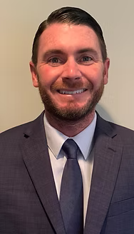 Man wearing a dark suit and tie, smiling at the camera. He has a short beard and hair, and is indoors.