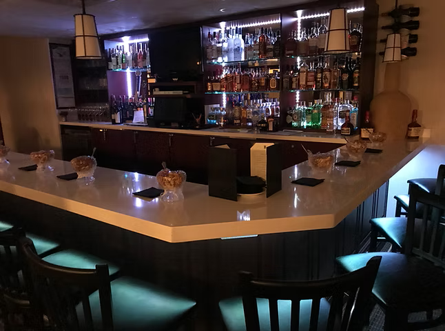 Bar with a white countertop, dark chairs, and shelves stocked with liquor bottles.