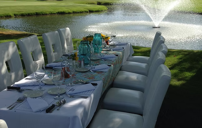 Outdoor table set for a meal with white chairs, next to a pond and fountain.