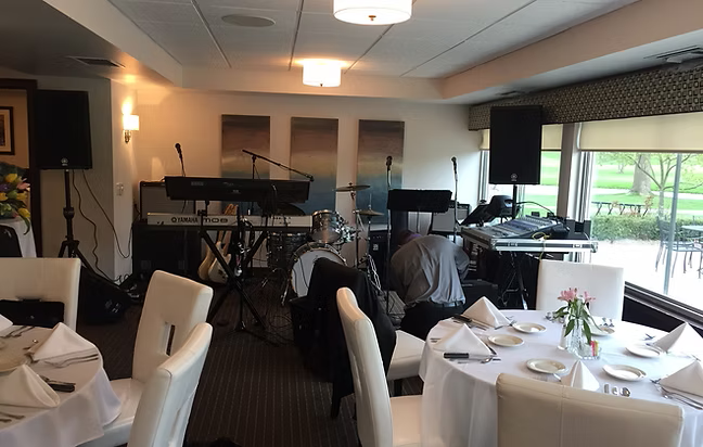 Restaurant with a band setting up: drums, keyboards, and speakers. Tables are set with white cloths and chairs.