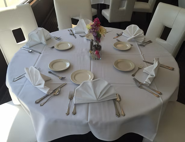 Round table set for six with white tablecloth, napkins, plates, silverware, and a flower arrangement.