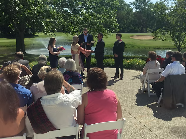 Wedding ceremony outdoors by a water feature; couple with officiant, guests seated in chairs.