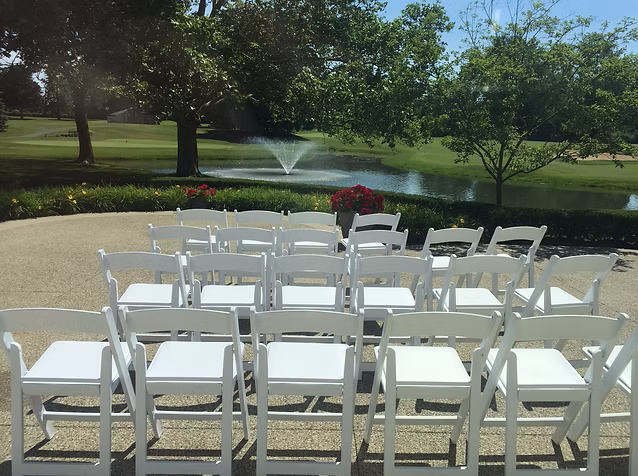 White chairs set up for an outdoor event, with a pond and trees in the background.