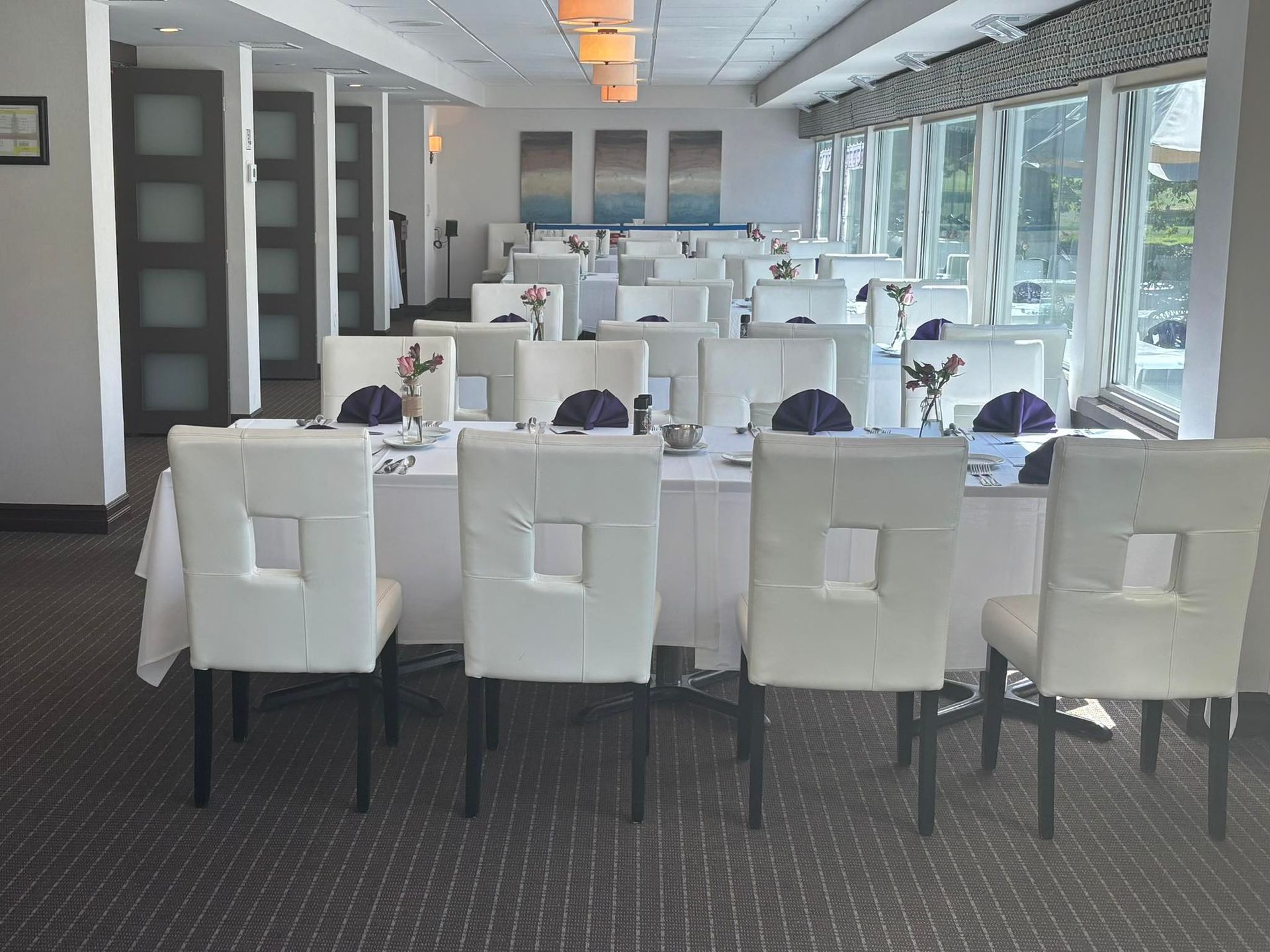 Elegant dining room with white tables, chairs, and table settings. Large windows and decorative panels.