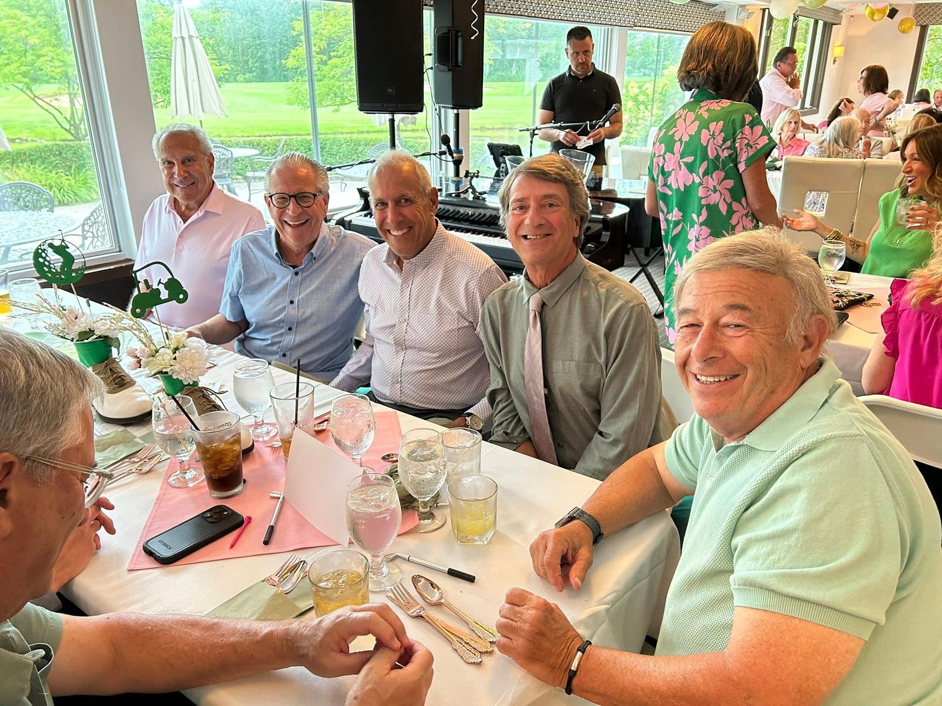 People seated at a table, smiling. Band playing in the background. White tablecloth with drinks and flowers.