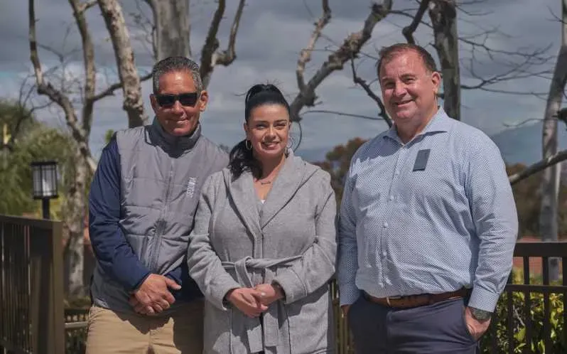 Three people stand outdoors. Woman in a gray coat flanked by two men. Trees and a deck are in the background.
