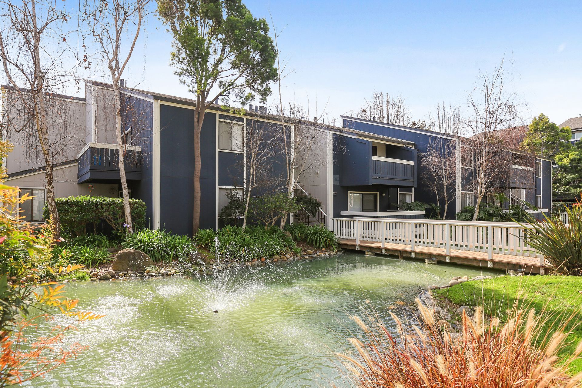 Apartment complex with a pond, fountain, and wooden bridge; blue and gray exterior.