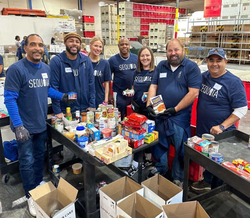 Volunteers wearing blue shirts sort food donations at a warehouse; various canned goods and boxes are on the table.