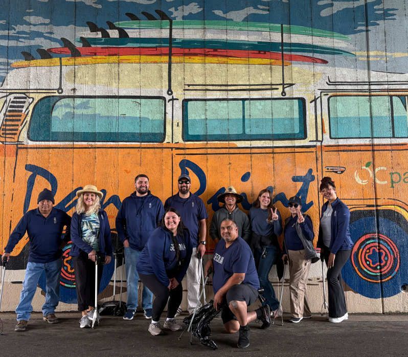 Group of people in front of a colorful mural of a bus with surfboards. They wear navy tops; some pose, smile.