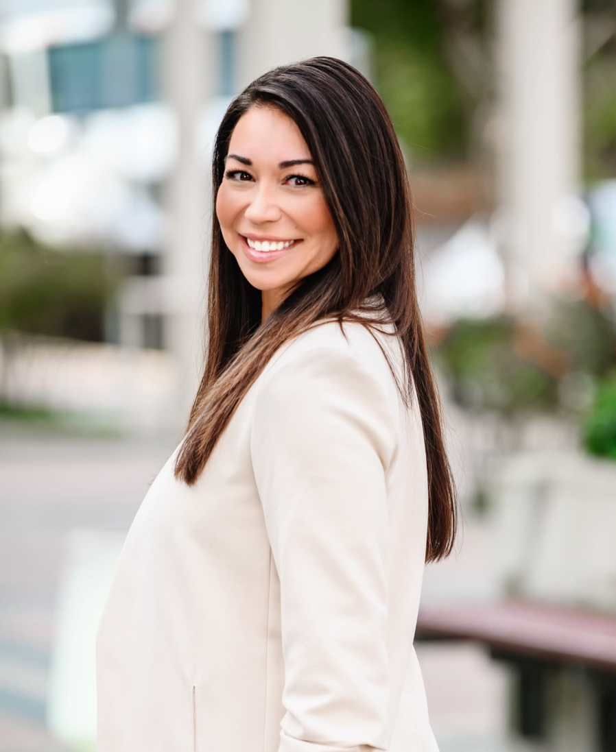 Woman with long dark hair, wearing a cream blazer, smiles towards the camera in an outdoor setting.