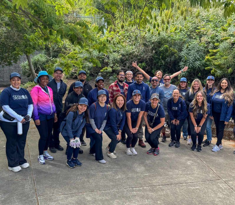 A group of people in navy blue shirts pose outside with trees in the background. Some are smiling, others are looking at the camera.