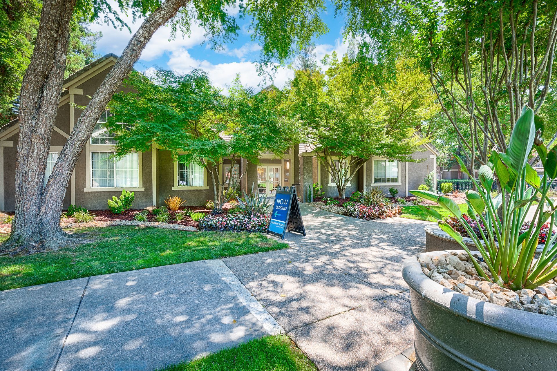 Lush green entrance to a building. Sidewalk leads to a doorway, flanked by trees and flowerbeds.