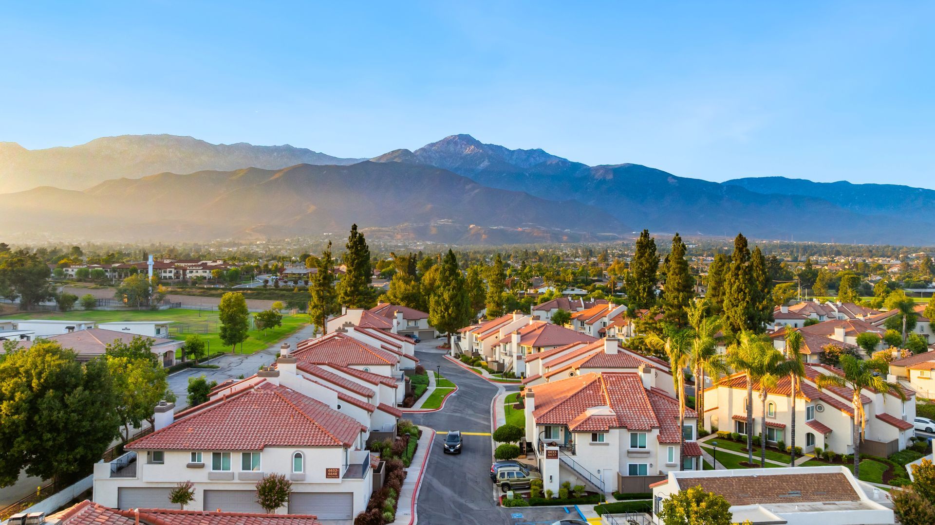 Row of townhouses with orange tiled roofs, trees, and mountains in the background under a blue sky.