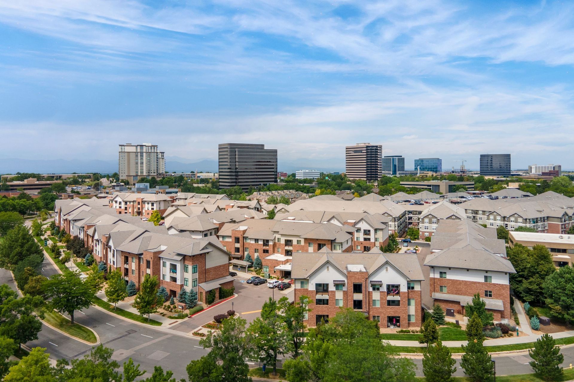 Aerial view of residential buildings with gray roofs, trees, and city buildings in the background under a blue sky.