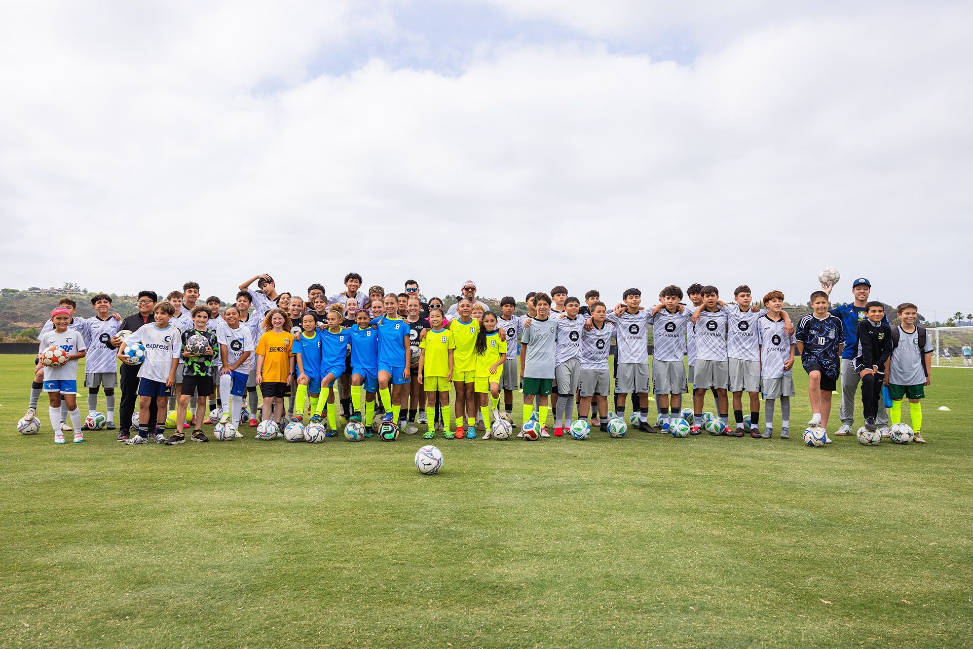 Large soccer team lined up on a grassy field, with two players in bright yellow at center and a ball in front.
