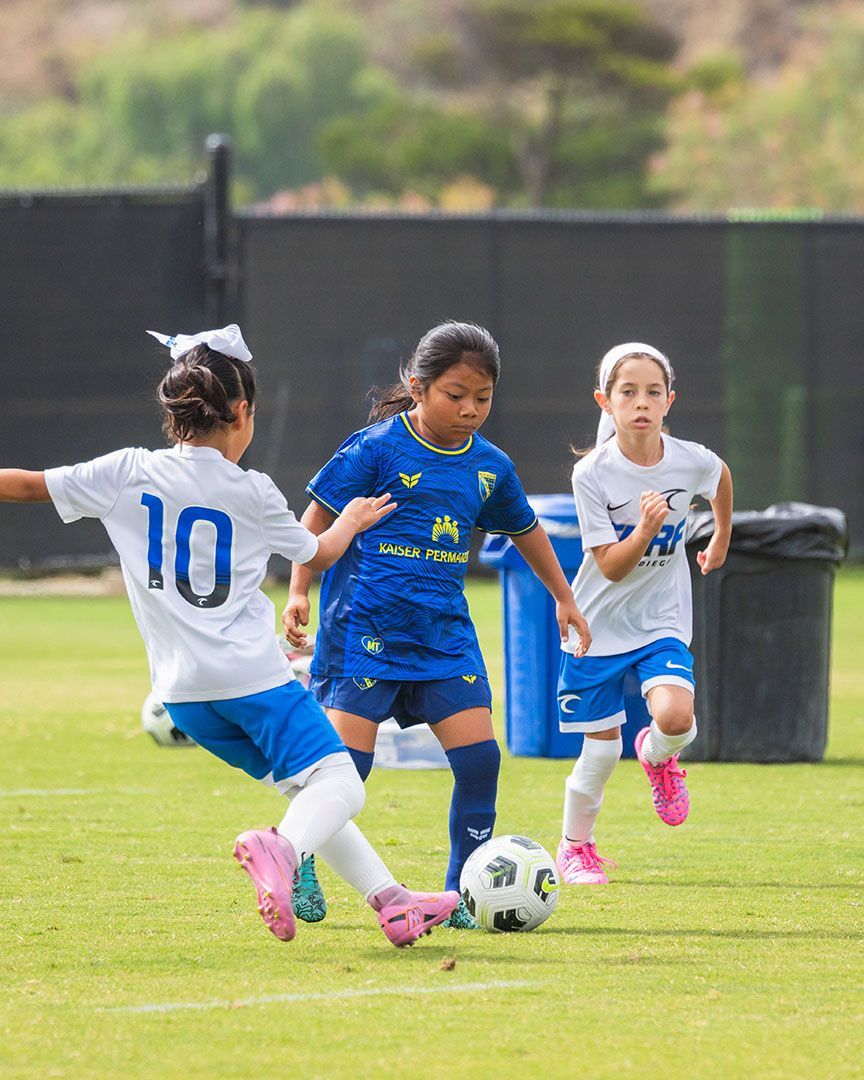 Girls playing soccer on a grassy field, with one in blue dribbling between two in white jerseys.